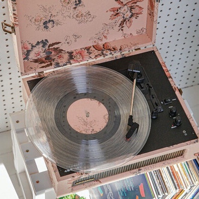 Gira-discos cor-de-rosa com flores e disco de vinil transparente a tocar, mesa branca, discos e plantas