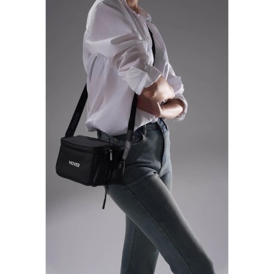 Bolsa preta com alça ao ombro, usada com roupa casual sobre fundo cinzento