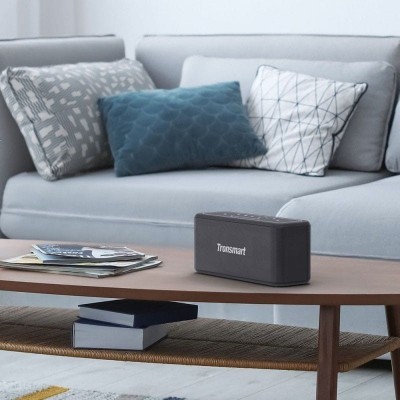 Coluna sonora Tronsmart preta sobre mesa de madeira, com sofá e almofadas ao fundo