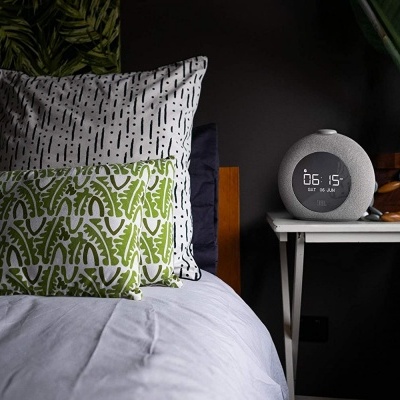 Relógio digital numa mesa ao lado da cama com almofadas e plantas