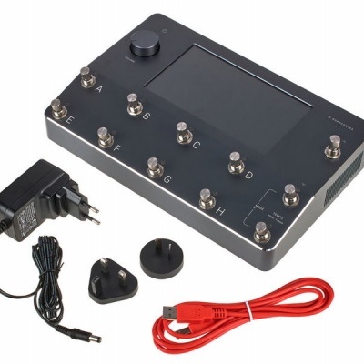 Aparelho preto com visor e doze interruptores etiquetados, adaptador de energia e cabo USB vermelho.