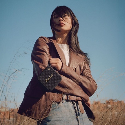 Mulher com casaco castanho e jeans segurando coluna portátil preta Marshall ao ar livre