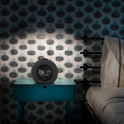 Relógio digital numa mesa de cabeceira azul junto a uma cama com almofada amarela e parede com padrão geométrico