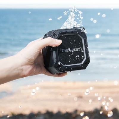 Coluna portátil preta Tronsmart com água a ser espirrada, com praia e mar ao fundo.