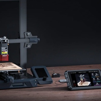 Impressora 3D preta com ecrã digital e tablet mostrando interface de impressão, sobre mesa de madeira