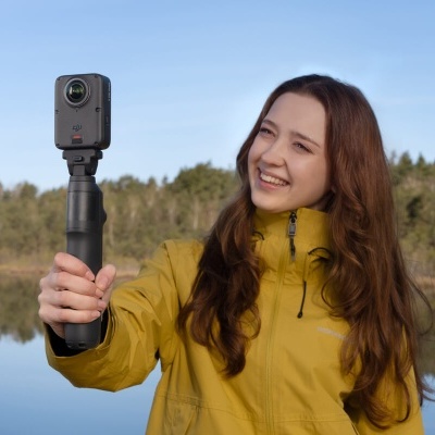 Mulher segurando câmara DJI preta de ação em suporte preto ao ar livre junto a lago