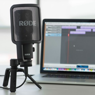 Microfone RØDE preto com filtro pop e tripé numa mesa junto a portátil com software de áudio