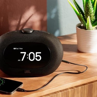 Rádio-relógio digital preto JBL com smartphone ligado e planta em vaso branco sobre móvel de madeira clara