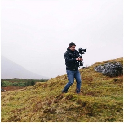 Homem com casaco preto e jeans a segurar uma câmara com estabilizador na natureza