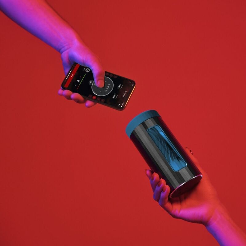 LELO F1S™ V3 – Masturbador Masculino Inteligente com App | Teal Mãos segurando smartphone com app e caixa cilíndrica preta com tampa azul em fundo vermelho.