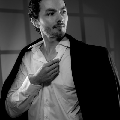 Homem de cabelo escuro com camisa branca e casaco escuro sobre os ombros em fotografia preto e branco