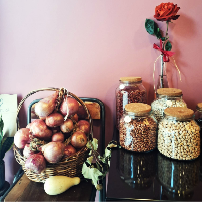 Vários frascos de vidro com feijão, cesto com cebolas e uma rosa vermelha num vaso em fundo rosa