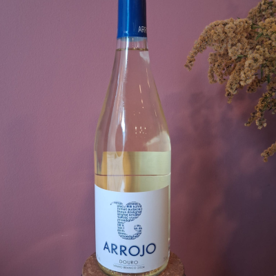 Garrafa de vinho branco com rótulo branco e tampa azul, perto de flores secas, contra parede rosa.