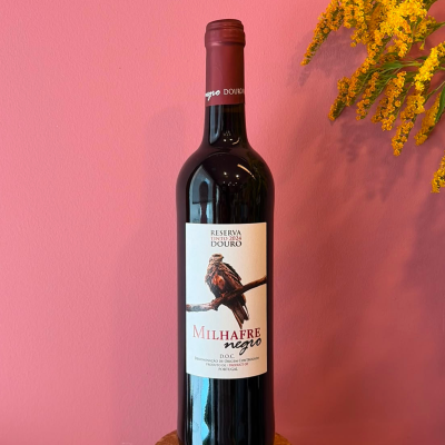 Garrafa de vinho tinto Milhafre Negro com rótulo branco e imagem de ave, contra fundo rosa e flores amarelas