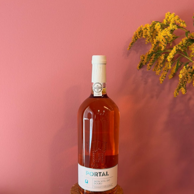 Garrafa de vinho rosé com rótulo branco numa superfície de cortiça contra parede cor-de-rosa com flores amarelas.
