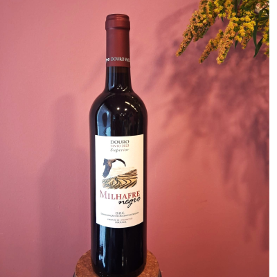 Garrafa de vinho tinto Milhafre negro com rótulo branco e flores amarelas contra parede rosa.