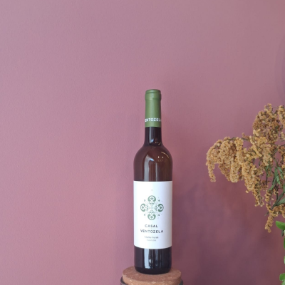 Garrafa de vinho CASAL VENTOZELA com rótulo branco e detalhes verdes sobre bases, parede rosa e flores secas de fundo