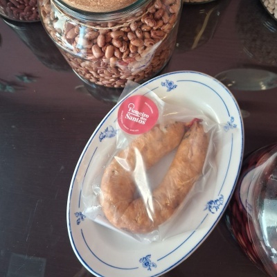 Linguiça embalada a vácuo com rótulo vermelho, prato branco com padrão azul, jarro de vidro com feijão castanho