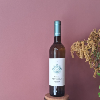 Garrafa de vinho branco Casal de VentoZela com rótulo branco e azul sobre suporte de cortiça em fundo rosa e ramo seco