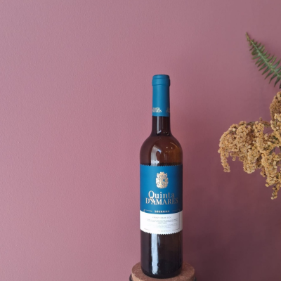 Garrafa de vinho Quinta DAmares com rótulo azul e branco e cápsula azul, junto a flores secas contra parede rosa