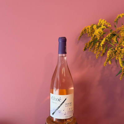 Garrafa de vinho rosé Oboé Superior Douro 2024 com rótulo branco e desenho de clarinete, em fundo rosa com flores amarelas.