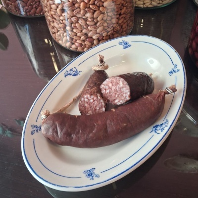 Linguiça preta cortada em prato branco com padrão azul e frascos de feijão ao fundo
