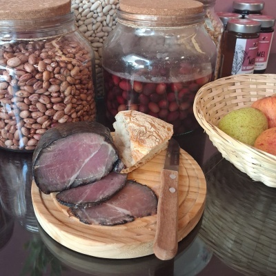 Tabua com carne curada e pão, frascos de vidro com feijão, azeitonas, mel e cesta com fruta