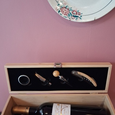 Caixa de madeira com garrafa de vinho e acessórios para vinho, parede rosa, prato decorativo pendurado.