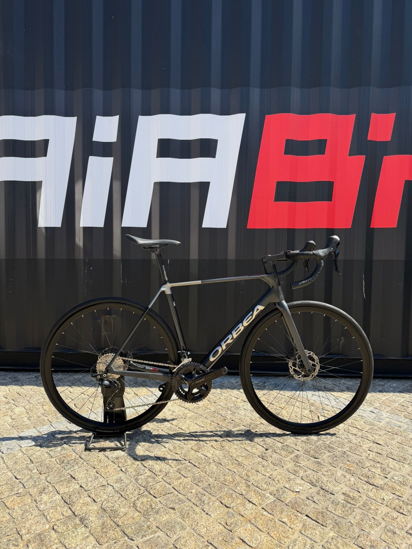 Orbea Orca Usada Preto/Cinza 53 Bicicleta de corrida preta Orbea apoiada em suporte com fundo preto, texto vermelho e branco.