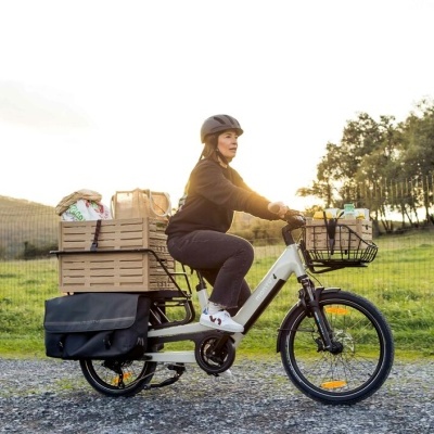Bicicleta elétrica branca com cesto e caixas de madeira carregadas na traseira, com passageira com capacete preto