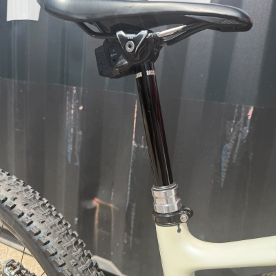 Sela preta de bicicleta com canote ROCKSHOX e quadro bege próximo a pneu