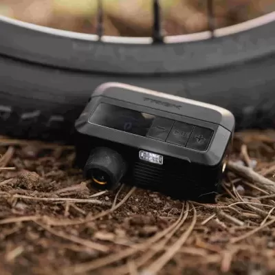 Dispositivo preto com visor digital e botões perto de pneu de bicicleta no chão com folhas secas
