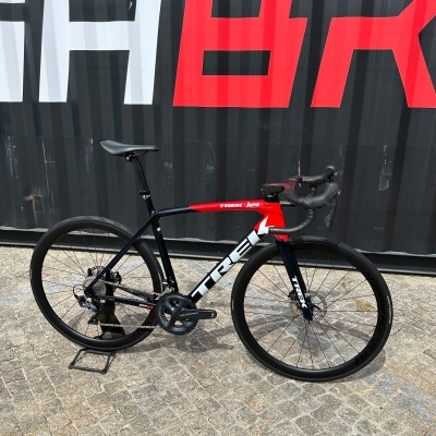 Bicicleta de estrada TREK preto, vermelho e branco encostada a um fundo com azulejos cinzentos