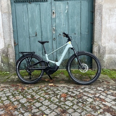 Bicicleta elétrica cinza claro com detalhes pretos encostada a uma porta verde antiga em chão de calçada portuguesa