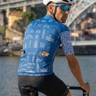 Ciclista com camisola azul de padrão e capacete branco junto a rio e ponte