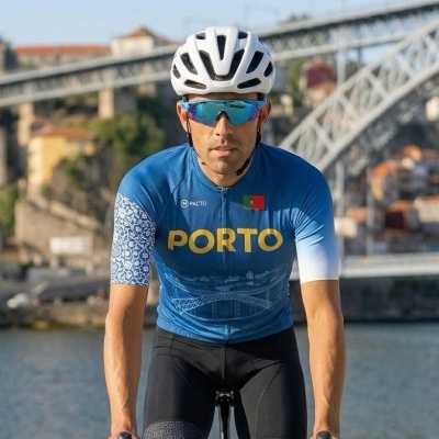 Ciclista com camisola azul de ciclismo PORTO, capacete e óculos, em fundo com ponte e rio.