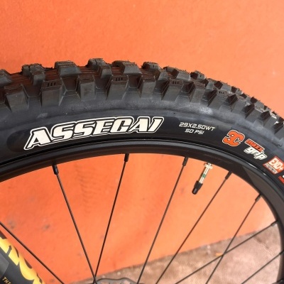 Pneu de bicicleta preto com texto ASSEGAI e especificações, montado numa roda com raios pretos contra fundo laranja.