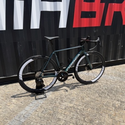 Bicicleta de estrada ORBEA azul escuro metálico com rodas brancas e pretas junto a contentor preto com letras brancas e vermelhas