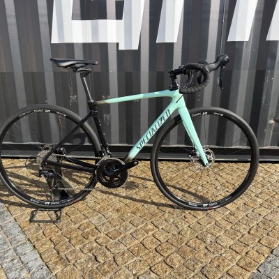 Bicicleta de estrada Specialized em degradê azul claro para preto, com rodas DT Swiss pretas, num suporte sobre piso em pedra