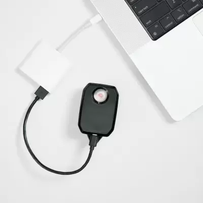 Adaptador USB preto ligado a computador portátil prateado com teclado preto num fundo branco.