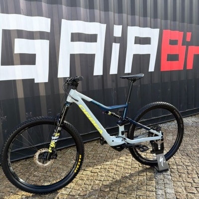 Bicicleta de montanha Orbea azul claro e azul escuro com pneus Maxxis e suspensão Fox apoiada no chão de calçada junto a um contentor cinzento