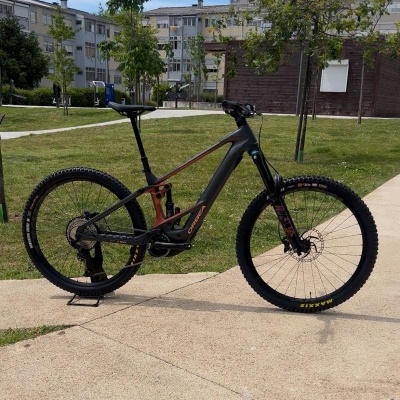 Bicicleta de montanha elétrica preta com detalhes vermelhos, pneus Maxxis, suspendida na calçada