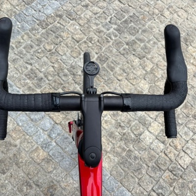 Guiador de bicicleta de estrada em carbono com fita preta, suporte para computador e quadro vermelho sobre pavimento de pedra cobblestone.