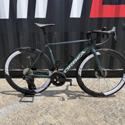 Bicicleta de estrada ORBEA azul esverdeada com rodas de pneus pretos e aros brancos, guiador curvo preto e selim preto encostada a estrutura metálica preta.