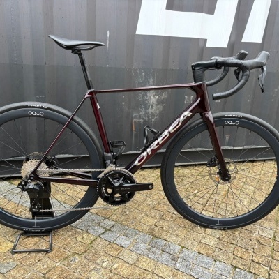 Bicicleta de estrada Orbea com quadro bordô e rodas pretas em pavê junto a uma parede.