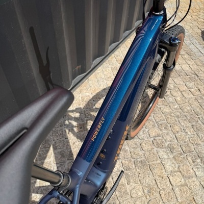 Bicicleta montanha azul metálica com pneus castanhos e textos no quadro junto a uma parede preta