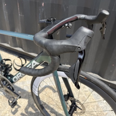 Bicicleta de estrada verde com componentes Shimano estacionada junto a parede preta