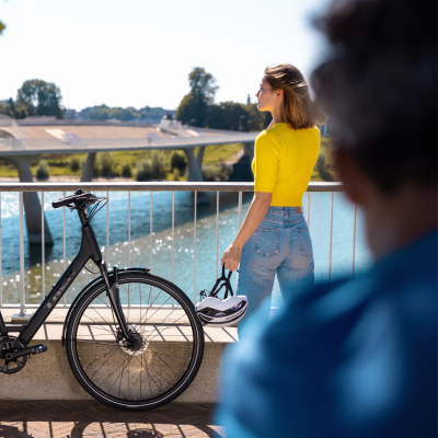 Mulher com camisola amarela e jeans junto a bicicleta preta junto a rio e ponte ao fundo