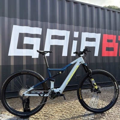 Bicicleta de montanha Orbea azul e cinzenta com pneus larges e suspensão dianteira, encostada a um contentor preto com as letras GAIA em branco e vermelho.