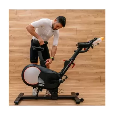 Homem ajusta assento de bicicleta estática preta com detalhes em vermelho e cinza, fundo de madeira clara